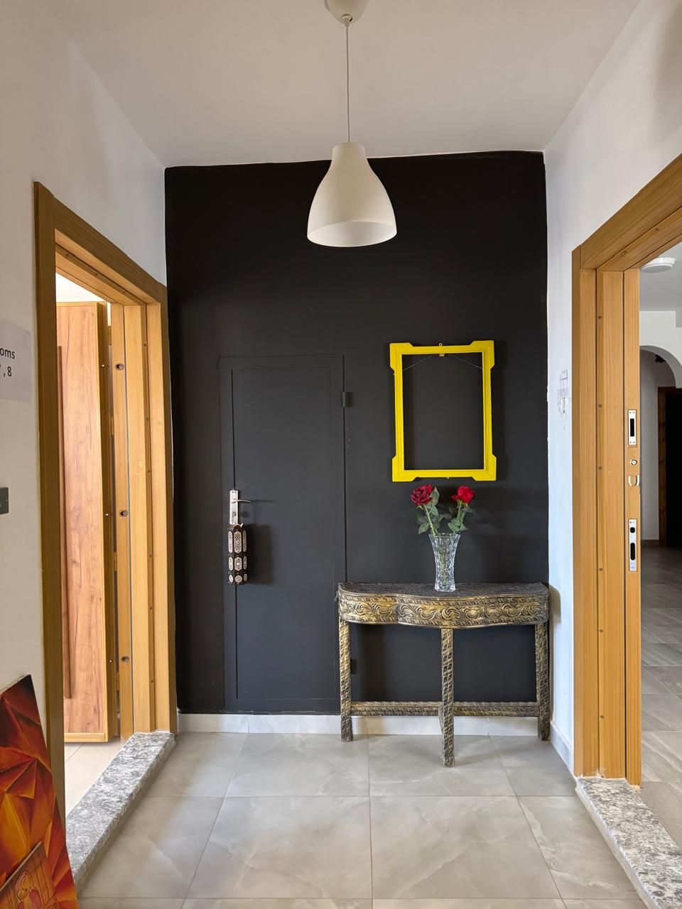 A hallway with a black door, a yellow-framed mirror, and a vase with red roses on a console table.