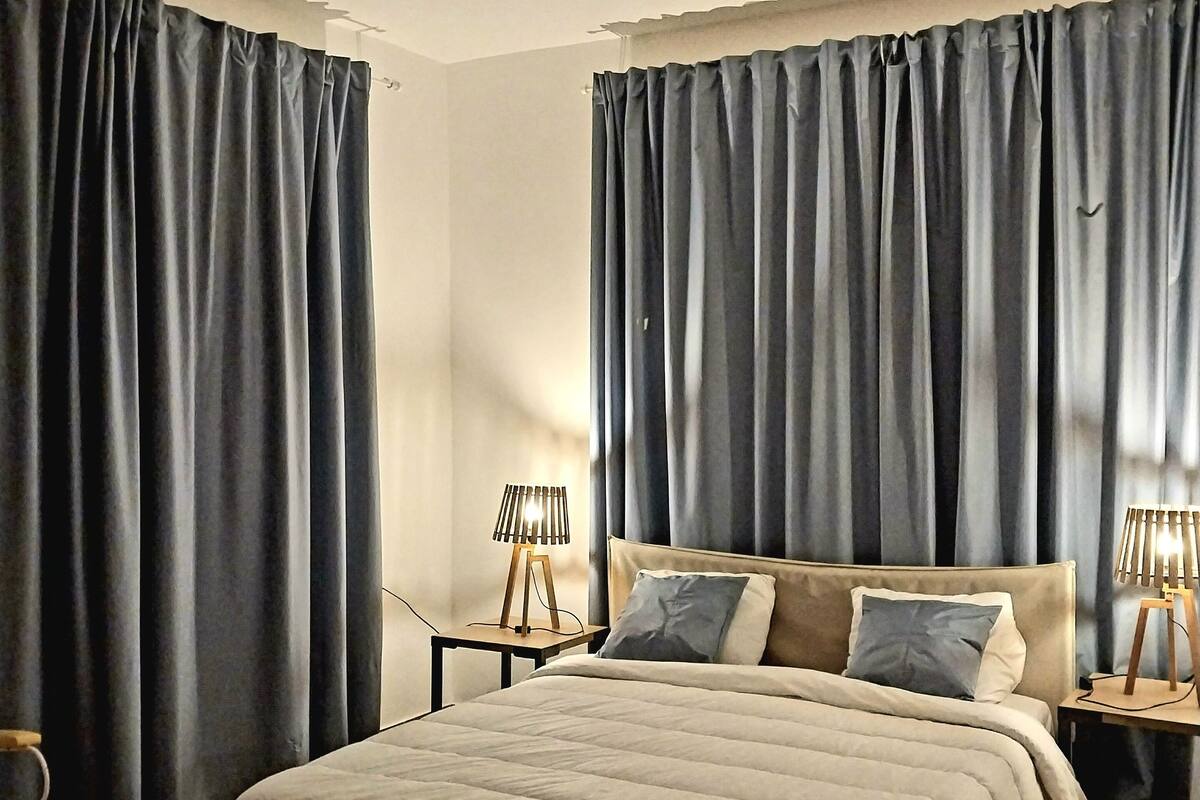 Ein Schlafzimmer mit einem Bett, zwei Nachttischen mit Lampen, dunklen Vorhängen und einem gemusterten Teppich.