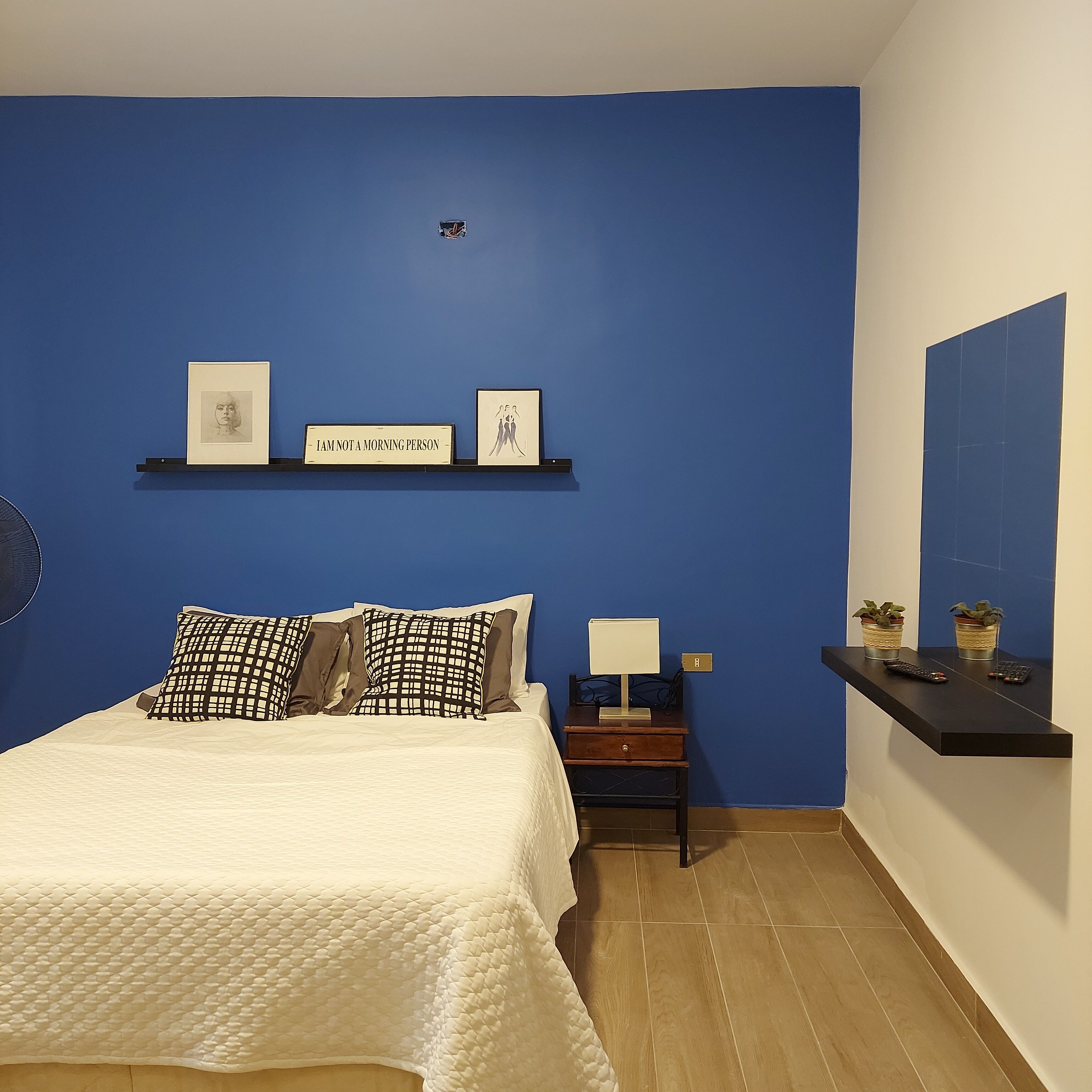 Camera da letto con un letto, cuscini, un comodino con una lampada, una mensola con delle foto e una sveglia a muro.