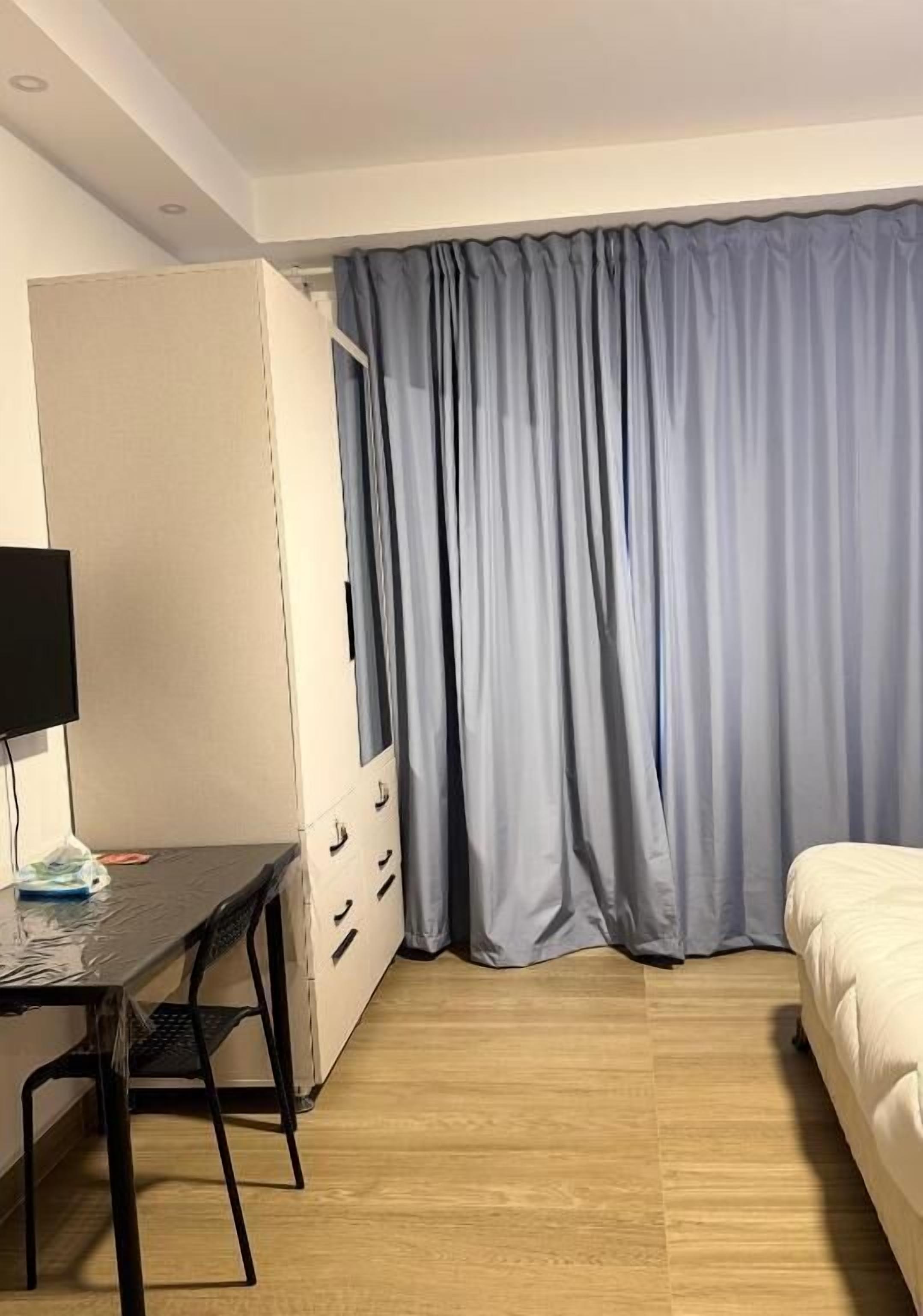 A hotel room with a bed, a desk with a chair, a wardrobe, and a large window with curtains.