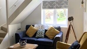 A cozy living room with a blue sofa, yellow armchair, and a wooden coffee table. A staircase leads to an upper level, and a tripod lamp is visible. - River and country views & woodburner at ‘The Nest’ (Burrowbridge)