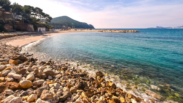 Una playa costera con agua turquesa cristalina, costa rocosa y un muelle distante.
