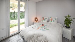 Un dormitorio con una cama grande, una mesita de noche con una lámpara, una planta en maceta y un ventanal corredero que da a un espacio exterior.