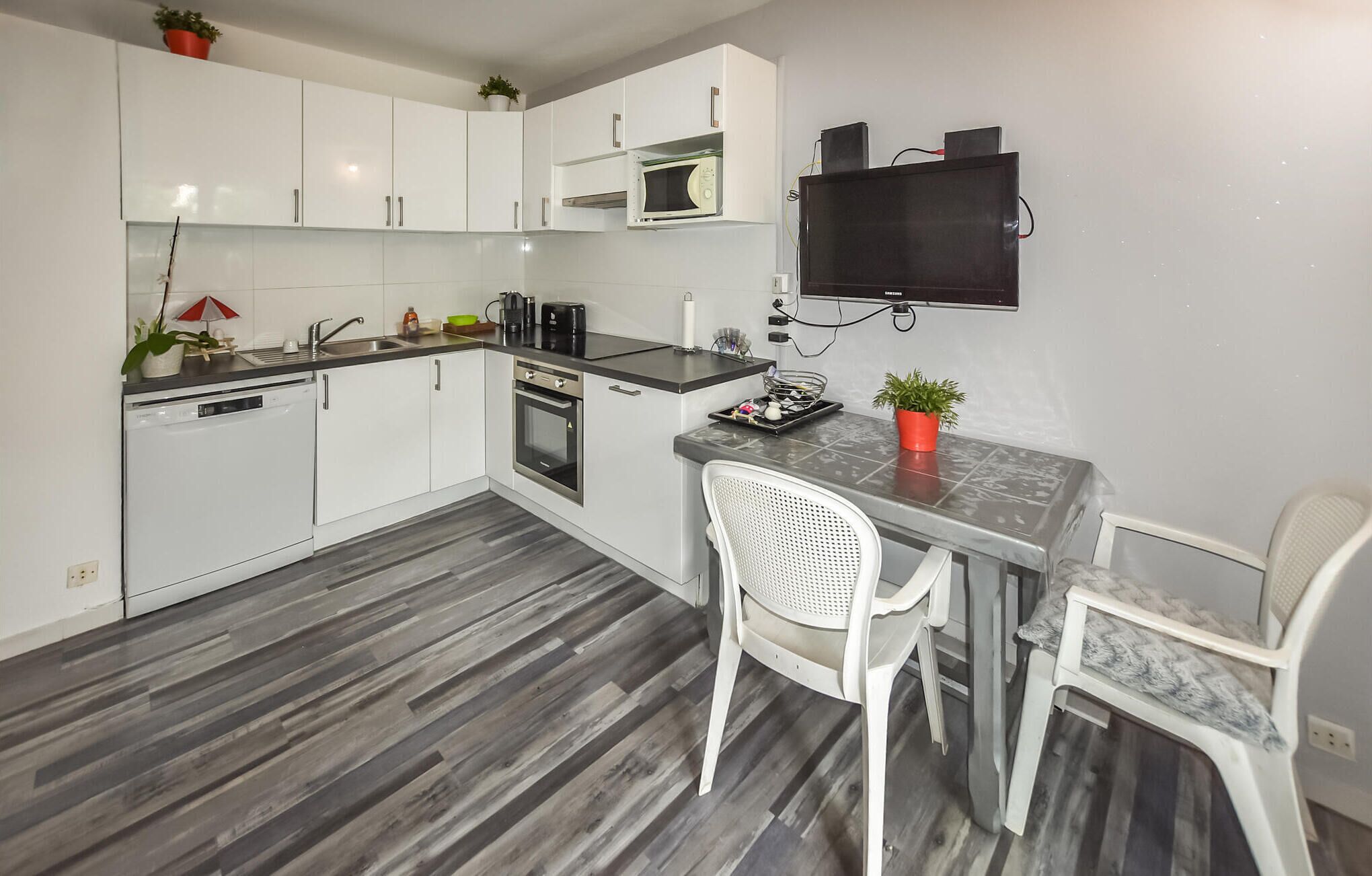 A compact kitchen with white cabinets, a microwave, a stove, and a TV mounted on the wall. There are two white chairs and a small table.