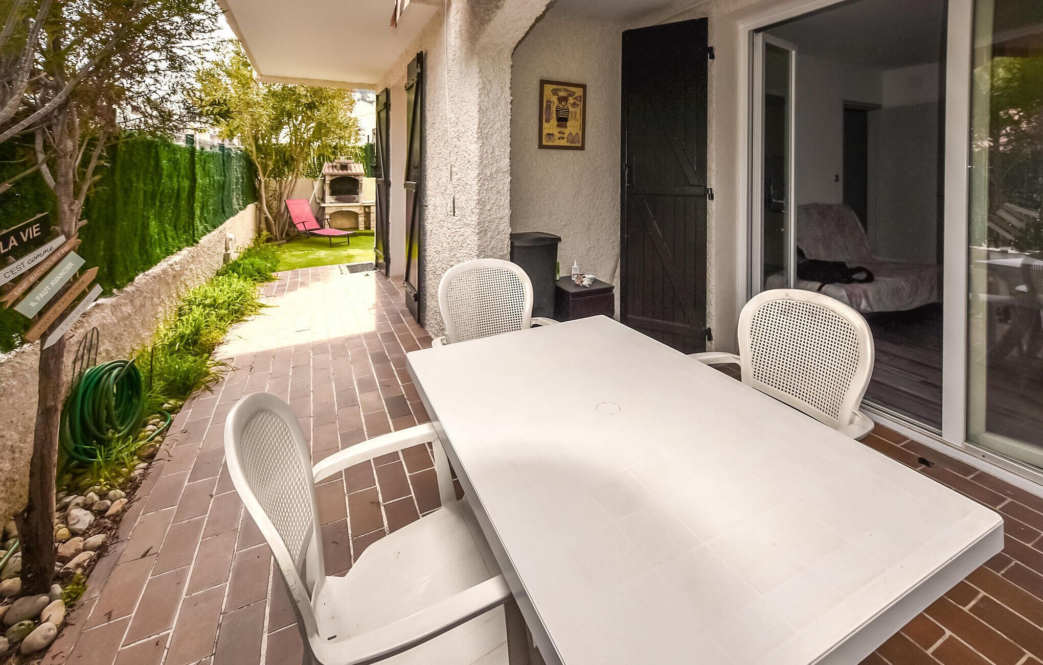 Un patio con mesa y sillas, una puerta corrediza de vidrio y un pequeño jardín con una silla roja y un horno de pizza.