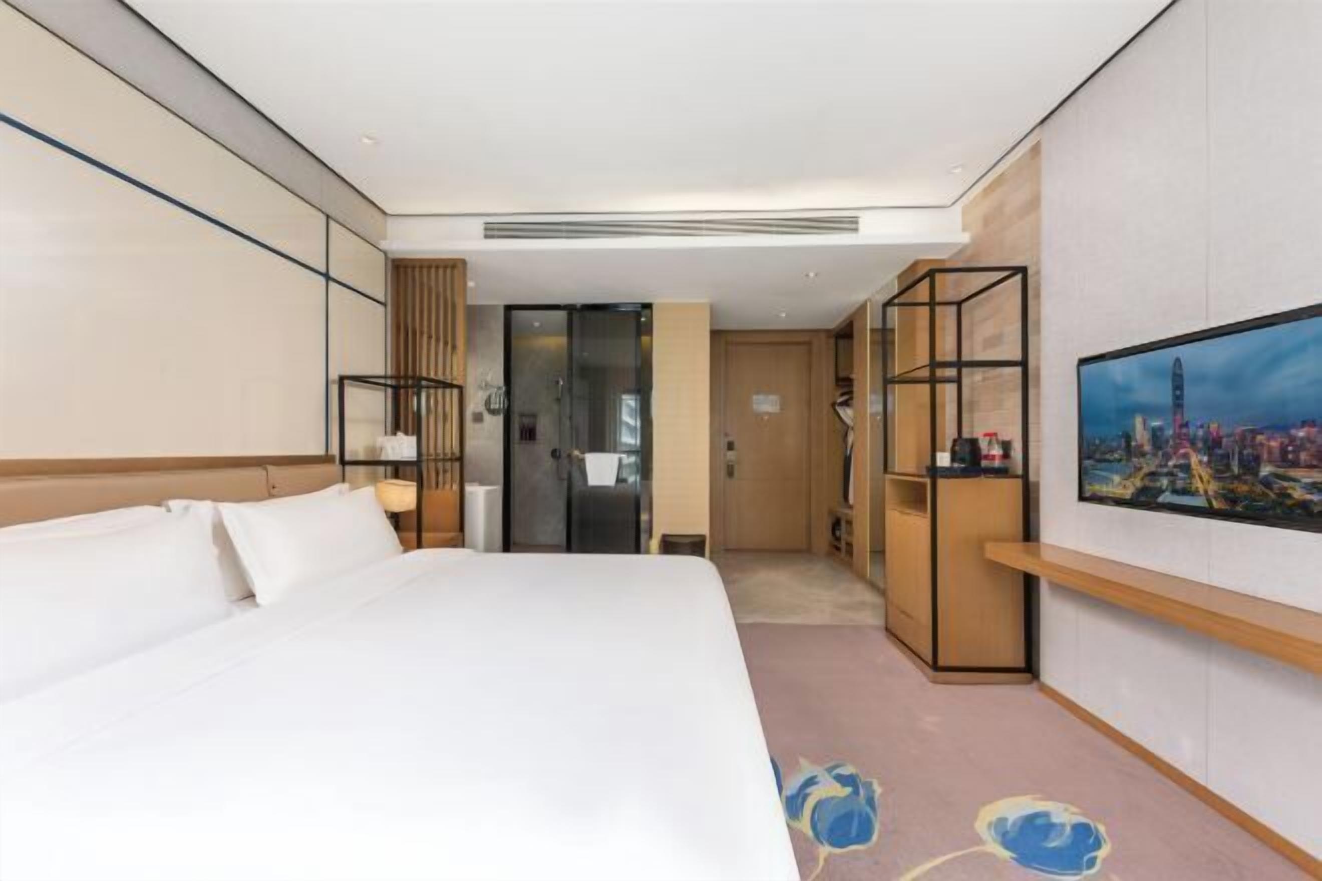 A hotel room with a large bed, a TV mounted on the wall, a wooden cabinet, and a glass door leading to a bathroom.
