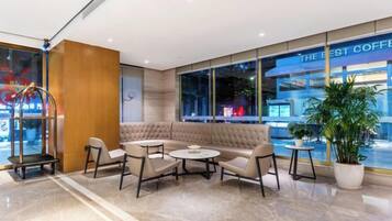 Un área lounge moderna con un sofá, sillas y mesas, un ventanal con vistas a una cafetería, y una planta en maceta.