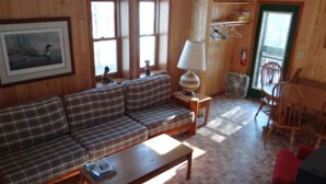 Ein Wohnzimmer mit Holzwänden, einer karierten Couch, einem kleinen Tisch und einer Lampe. Es sind Fenster, ein Sessel und eine Tür zu sehen.