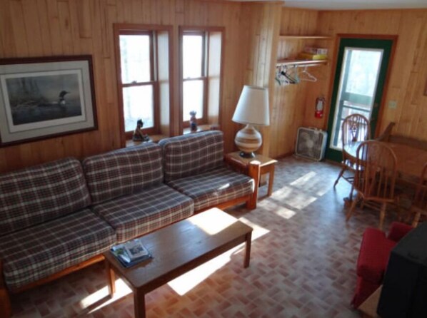 A wooden-paneled living room with a plaid sofa, a small table, and a lamp. There are windows, a chair, and a door visible. - The Bunk House (Stone Lake)