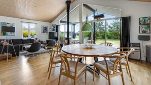 Uma sala ampla com uma mesa redonda de madeira e cadeiras, uma lareira e janelas grandes com vista para o exterior.