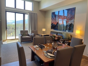 A dining area with a wooden table set for four, surrounded by comfortable chairs. A large window offers a view of a mountainous landscape. - Grand Colorado, 3 BD, master & 2 suites. Luxury Resort, steps to lifts & gondola (Breckenridge)