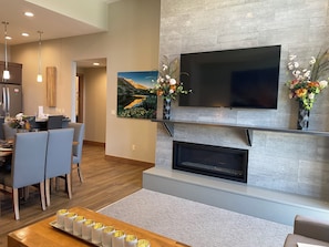 A modern living room with a flat-screen TV mounted above a fireplace, a dining area with chairs and a table, and a hallway leading to another room. - Grand Colorado, 3 BD, master & 2 suites. Luxury Resort, steps to lifts & gondola (Breckenridge)