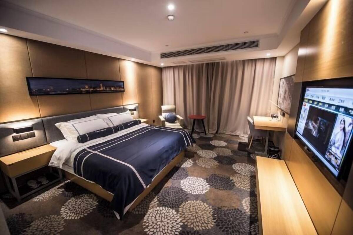 A modern hotel room with a large bed, a desk, a television, and a cityscape view.