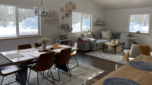 Ett rymligt vardagsrum med matplats, stora fönster med utsikt över snön, en takfläkt och en mysig öppen spis.