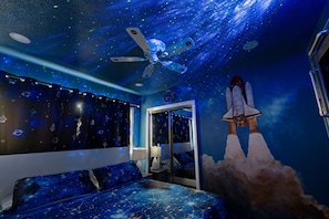 Une chambre avec un plafond décoré sur le thème de l’espace, un lit recouvert d’une couette à motifs galactiques et une fresque murale représentant le lancement d’une navette spatiale.