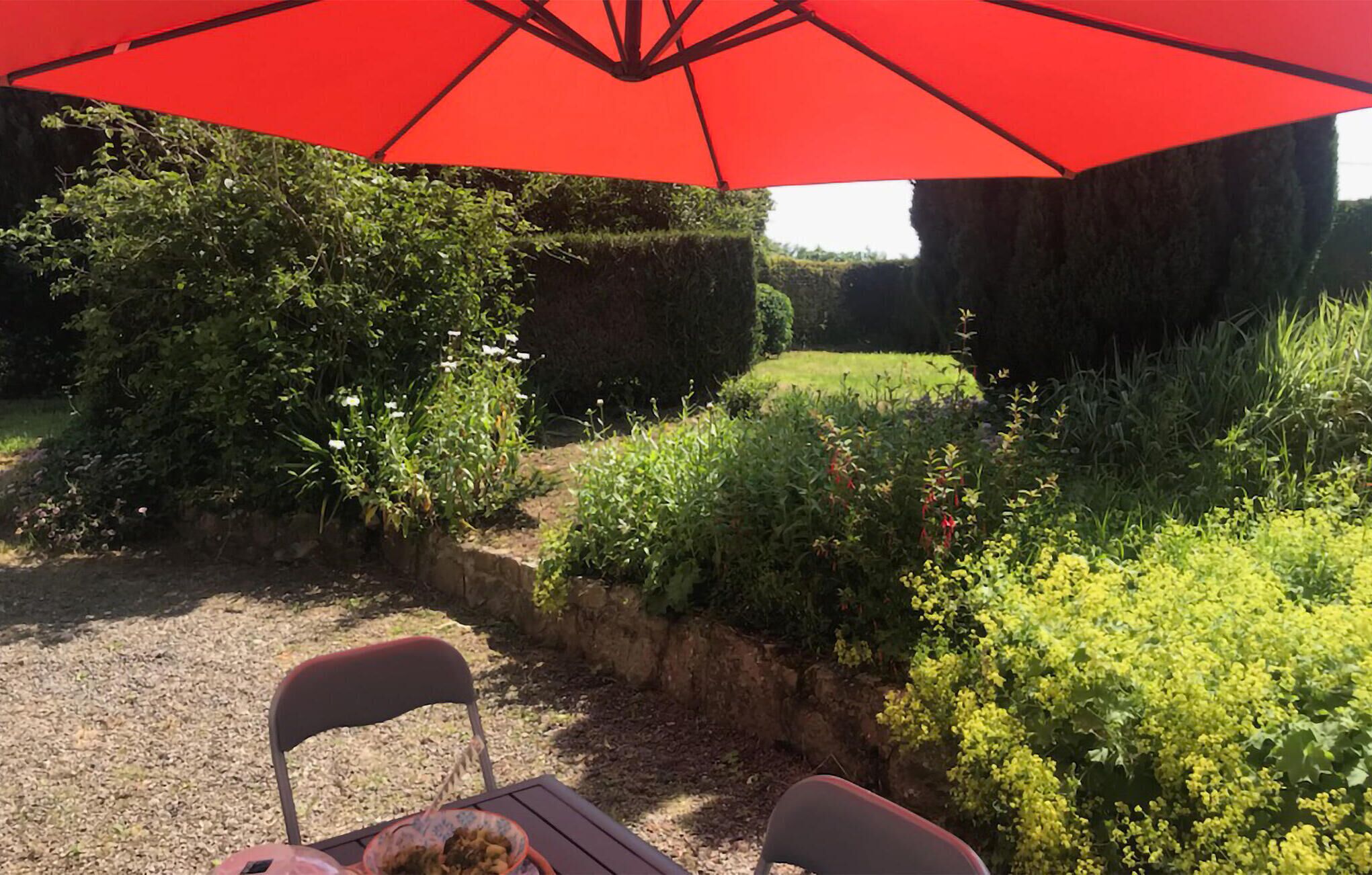 En have med et bord og stole under en rød parasol, omgivet af forskellige planter og hække.