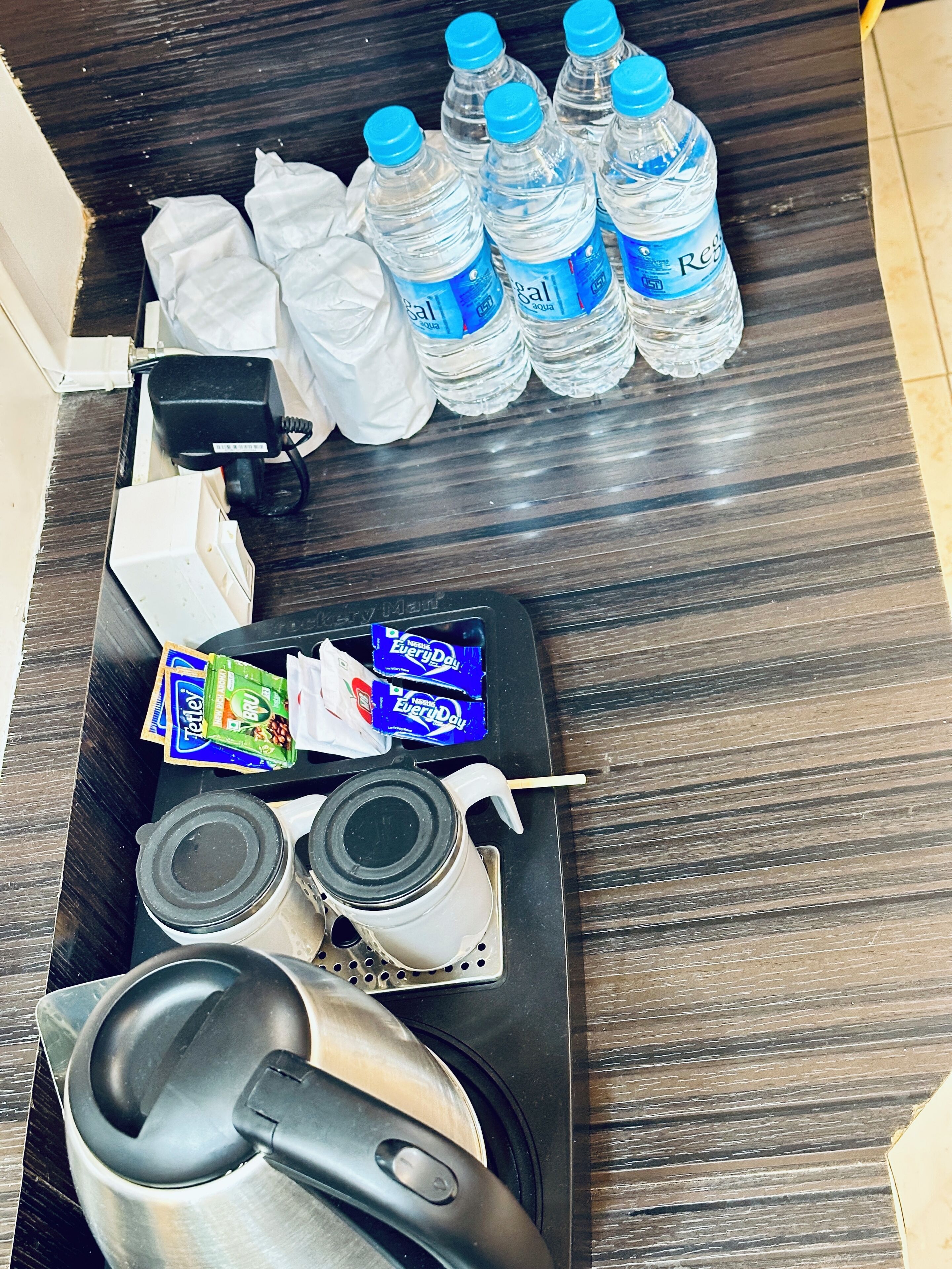 A hotel room with a wooden floor, a black tray with a coffee maker, two cups, a remote control, a pair of slippers, a tissue box, a water bottle, and a packet of snacks.
