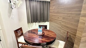 A dining area with a round wooden table, four chairs, a wall-mounted fan, and a window with curtains. - Saibaba International Hotel (Rahata)