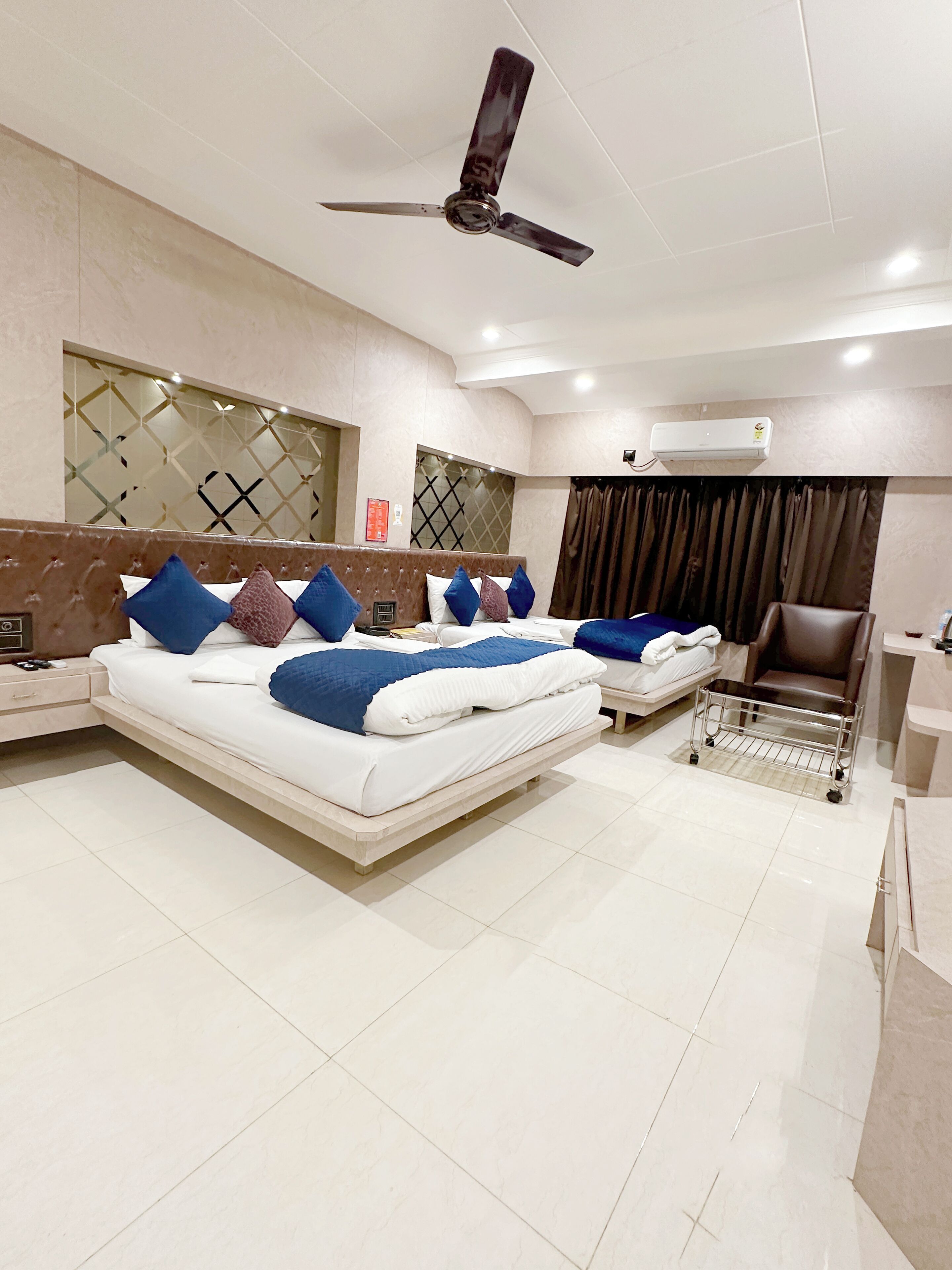 A modern hotel room with a large bed, a second smaller bed, a ceiling fan, an air conditioning unit, a chair, and a small table.