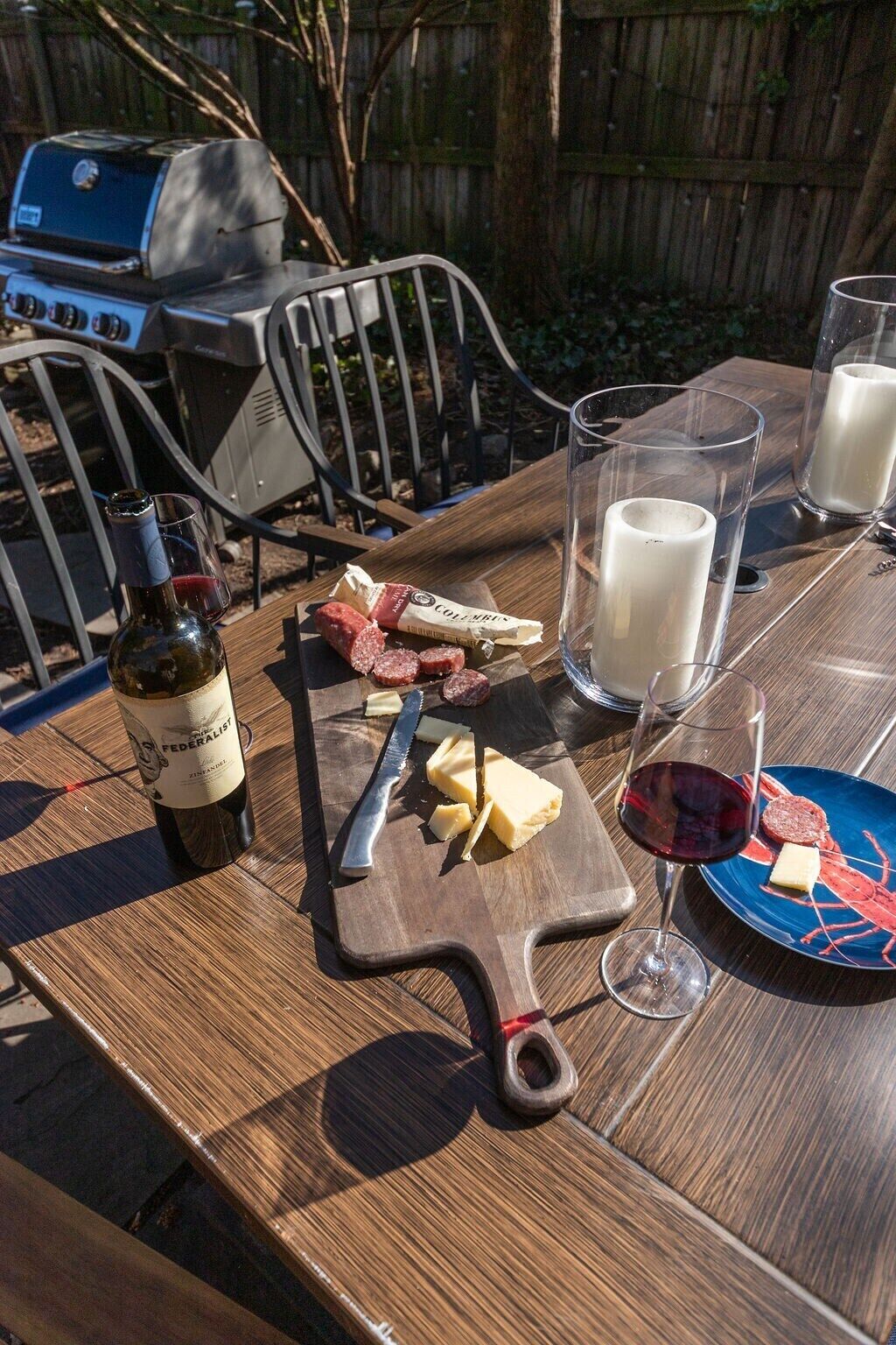 Een houten tafel met een fles wijn, kaas en een mes. Ook staan er melkglazen en een bord met eten op tafel.