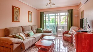 A living room with a sofa, a coffee table, and a television. There is a balcony with a chair and a table.