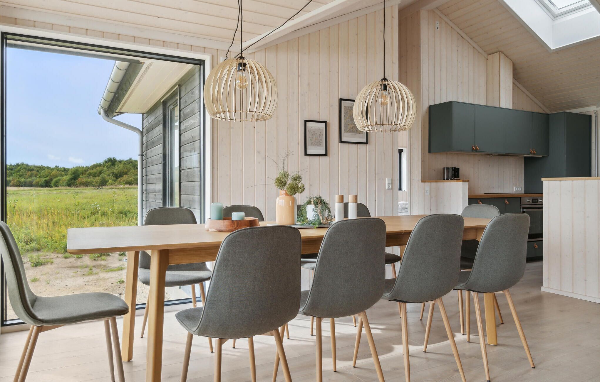 Uma sala de jantar moderna com mesa de madeira e cadeiras cinzentas, uma cozinha com armários escuros e vista para uma paisagem verde através de uma janela grande.