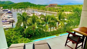 A balcony with two lounge chairs, a small table, and a view of a marina with boats and a building in the distance. - Relax in front of Ixtapa's beautiful Marina. (Ixtapa Zihuatanejo)