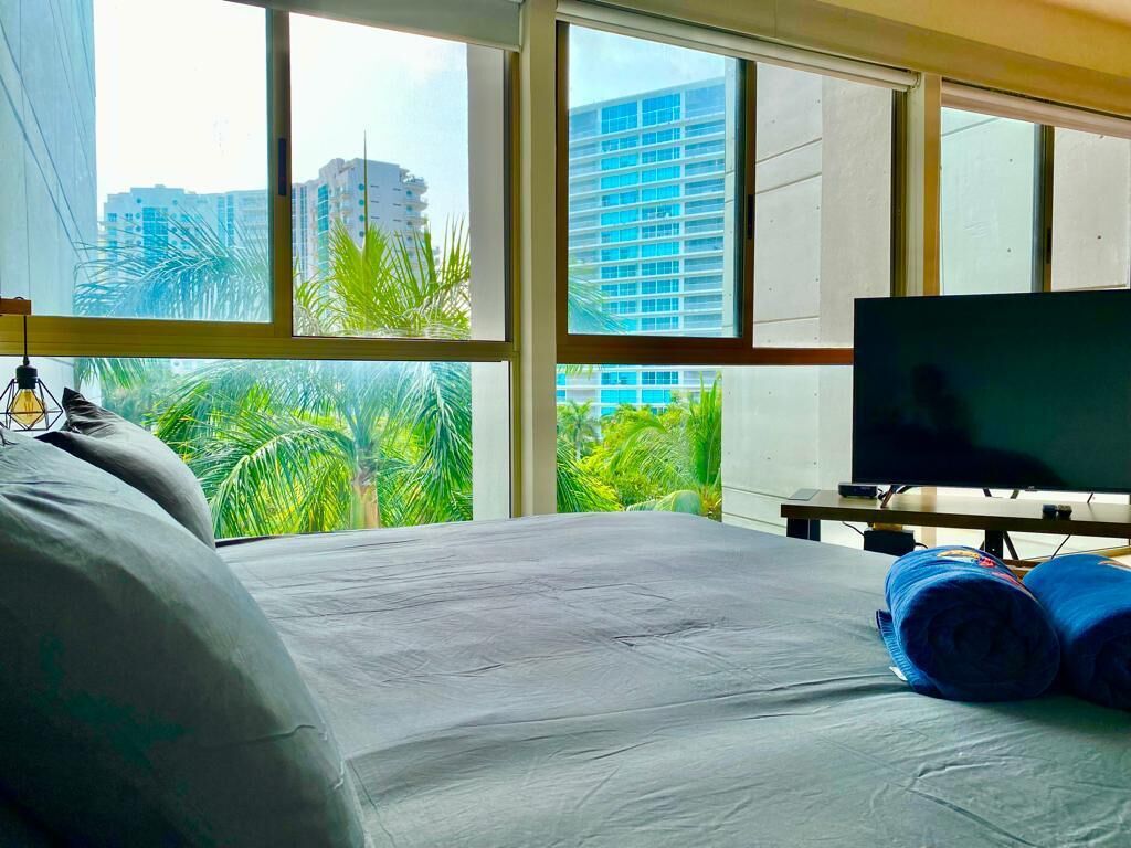 Une chambre d’hôtel avec un lit, une télévision et une fenêtre donnant sur des palmiers et des immeubles.
