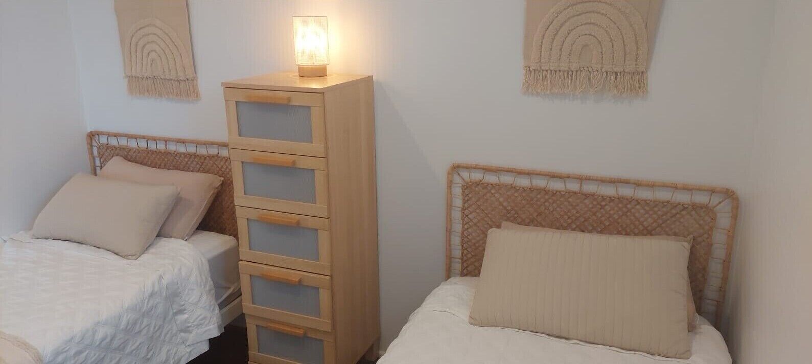 A bedroom with two single beds, a wooden nightstand with drawers, and a wall-mounted lamp.