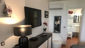 Quarto moderno com uma televisão de tela plana grande, luminária de cabeceira, uma pequena planta em vaso, uma cadeira branca e banheiro visível com pia e espelho.