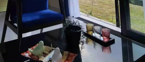 Un salon moderne avec un fauteuil bleu, une table basse en verre agrémentée d’objets décoratifs et une vue sur un paysage verdoyant, visible à travers de grandes fenêtres.