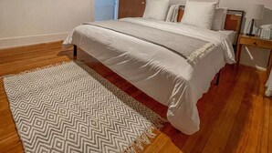 A bedroom with a wooden ceiling, a bed with white bedding, a patterned rug, and three framed pictures on the wall.