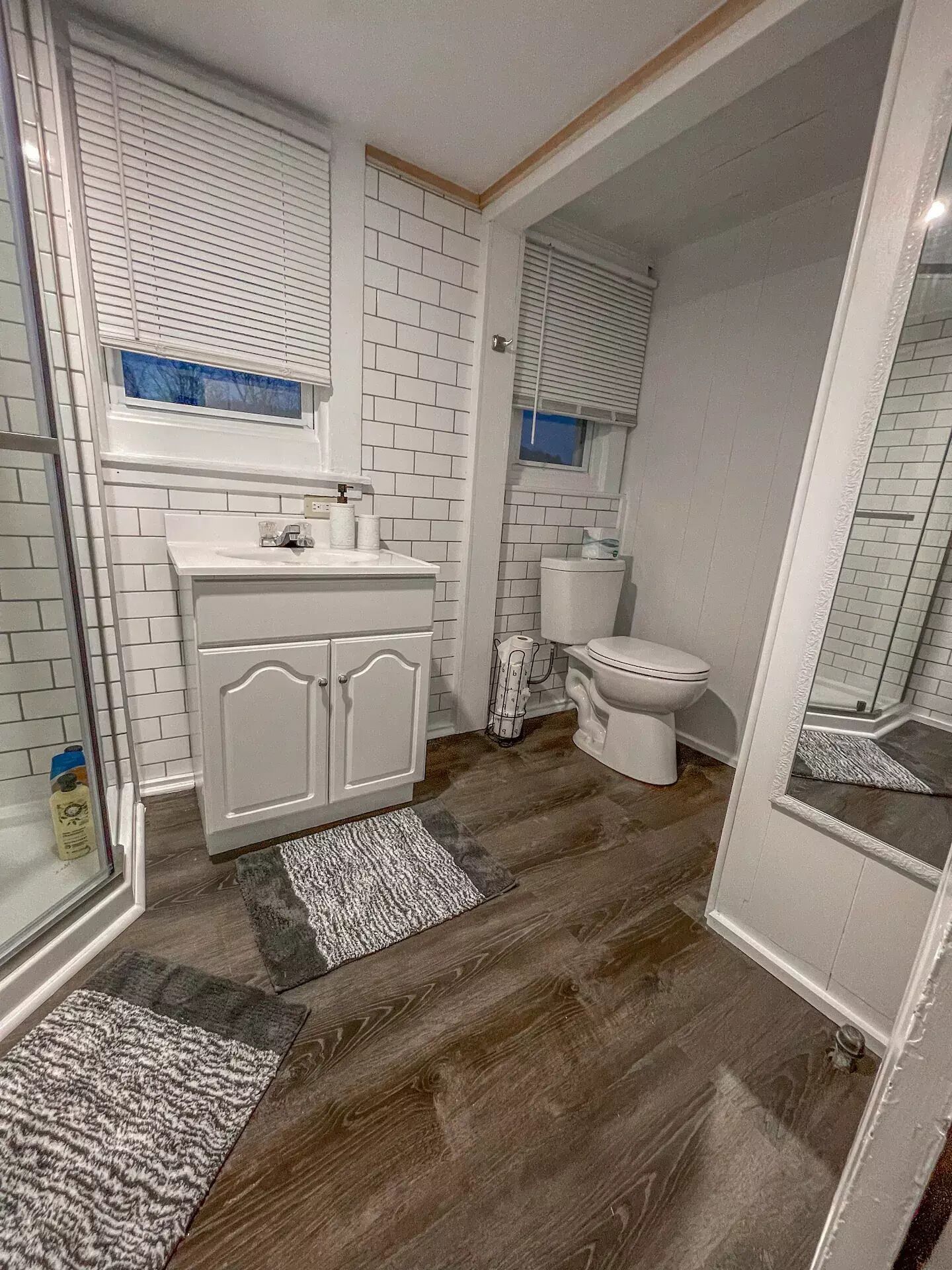 A bathroom with white tiled walls, a white vanity with a mirror, a toilet, and a shower area.