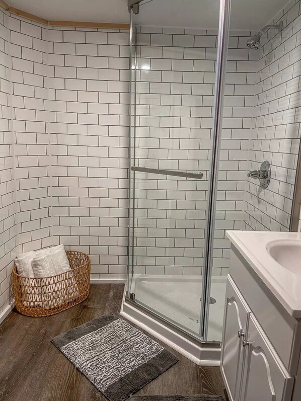 Une salle de bain moderne avec des carreaux de métro blancs, une douche en verre, une baignoire blanche et un panier en osier contenant des coussins.