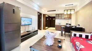 Ein modernes Hotelzimmer mit einem großen Flachbildfernseher, einem Kühlschrank, einem Esstisch mit Blumenstrauß, einer roten Couch und einer Küche im Hintergrund.