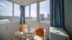 Ein Zimmer mit großem Fenster, einem Sessel, einem kleinen Tisch und einer Waschmaschine.