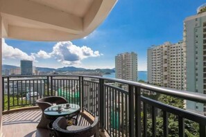 Un balcón con mesa y sillas, con vista a un paisaje urbano que incluye edificios altos y una masa de agua.