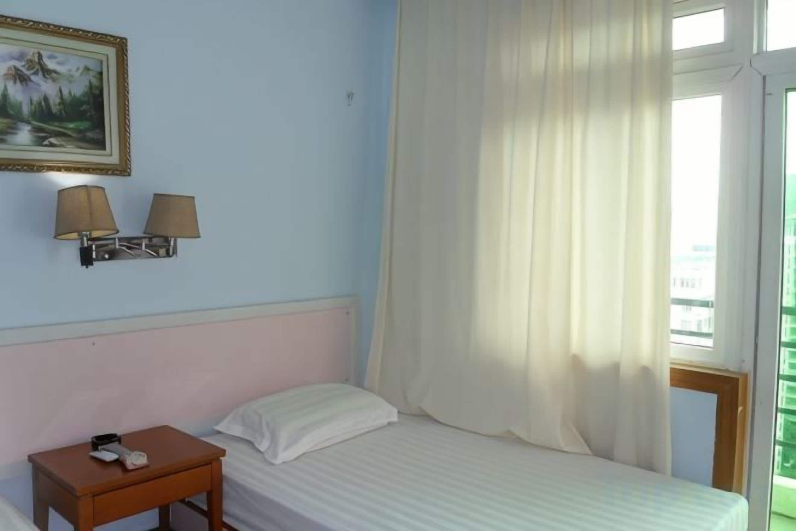 A hotel room with a bed, a nightstand, two wall-mounted lamps, a painting, and a window with curtains.