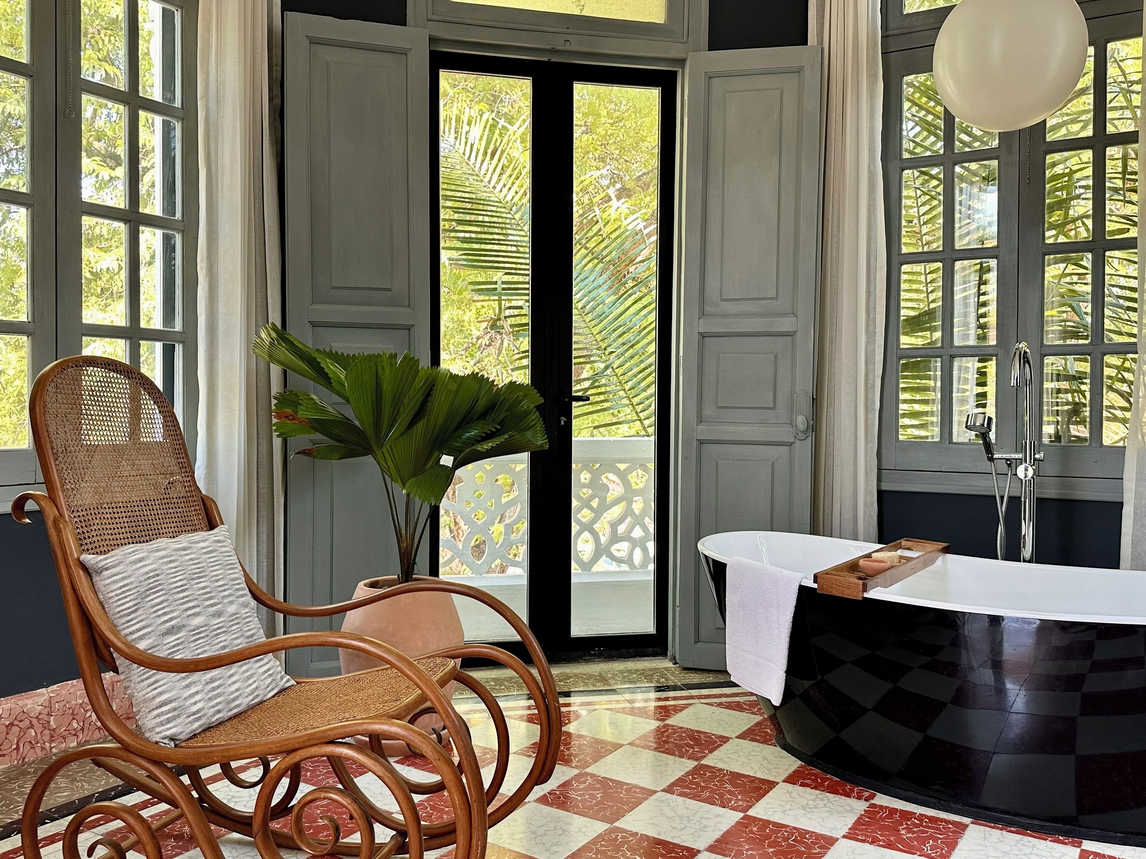 A room with a checkered floor, a wicker chair with a cushion, a potted plant, a bathtub, and a window with a view of greenery.
