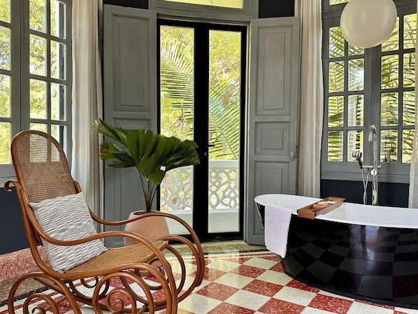 A room with a checkered floor, a wicker chair with a cushion, a potted plant, a bathtub, and a window with a view of greenery.