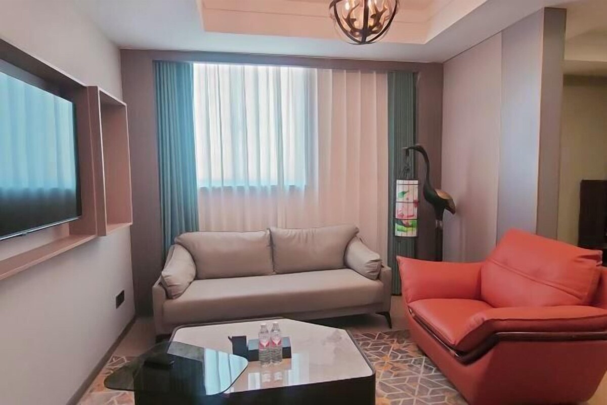 A living room with a sofa, a red chair, a coffee table with bottles, a TV, and a window with curtains.