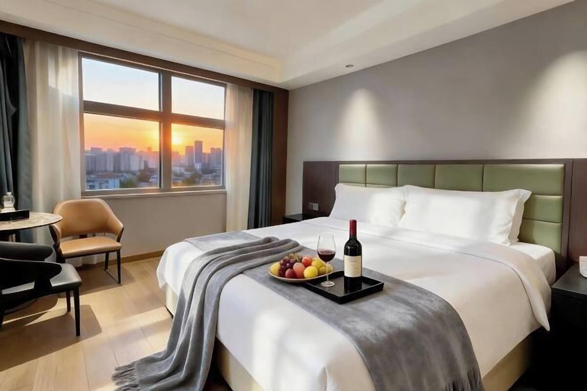 A hotel room with a bed, a dining table, a chair, a tray with fruit and wine, and a city view.