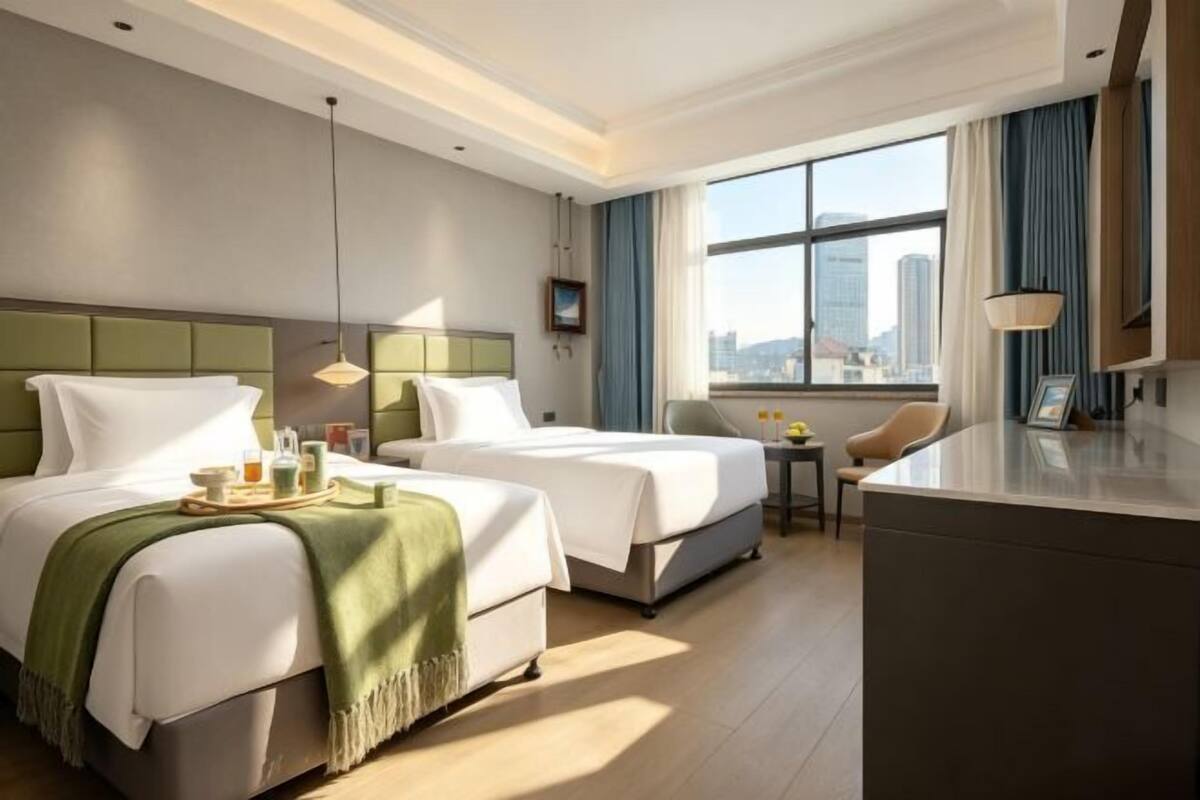 A hotel room with two beds, a desk, a chair, a small table, and a large window with a city view.