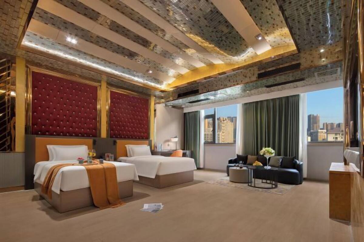 A hotel room with a large bed, two smaller beds, a sofa, and a coffee table. The room has a city view and a patterned ceiling.