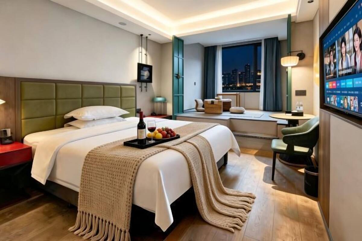 A hotel room with a bed, bedside table, TV, and a window with a city view.