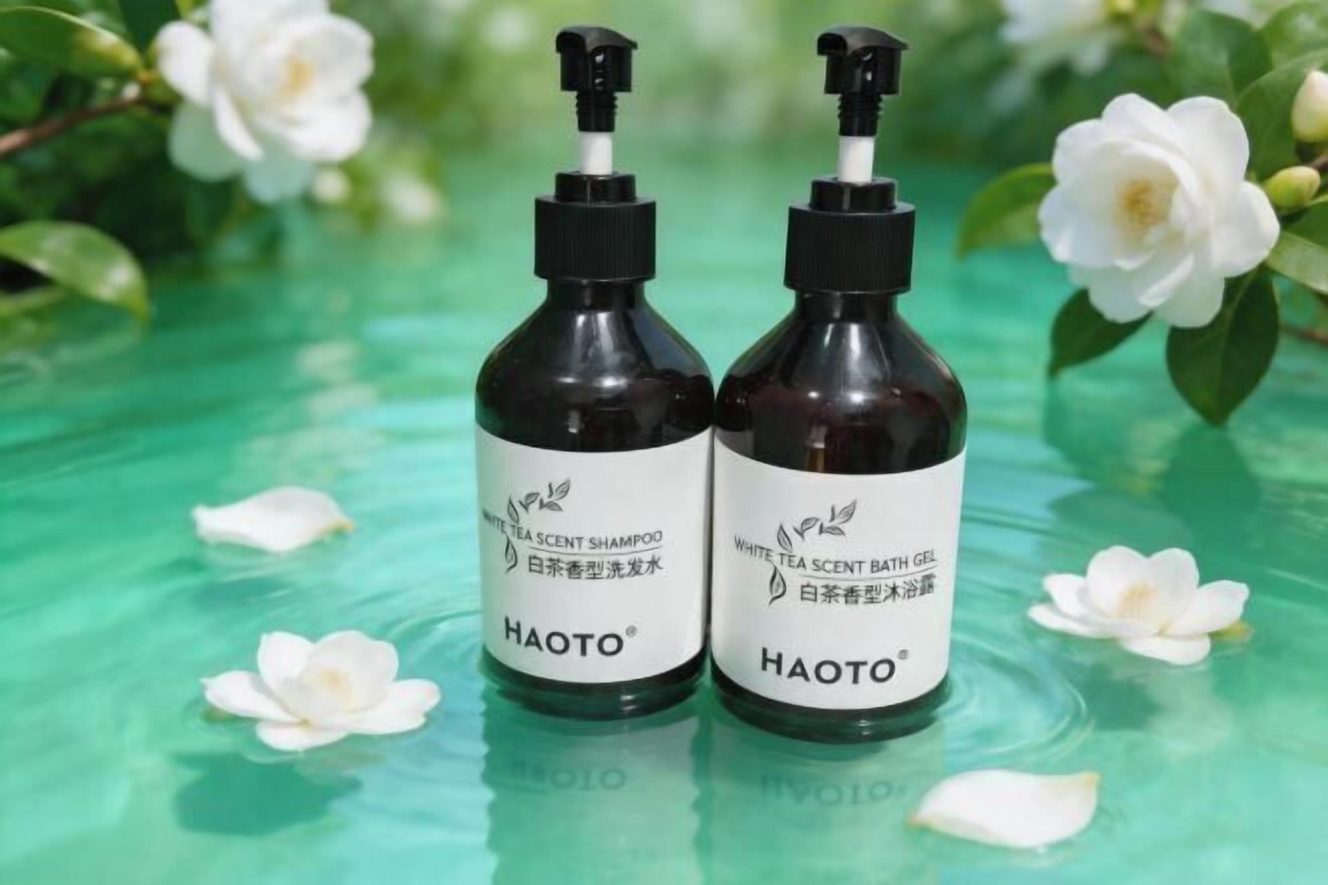Two bottles of Haoto white tea scent shampoo and bath gel on a reflective surface with white flowers.