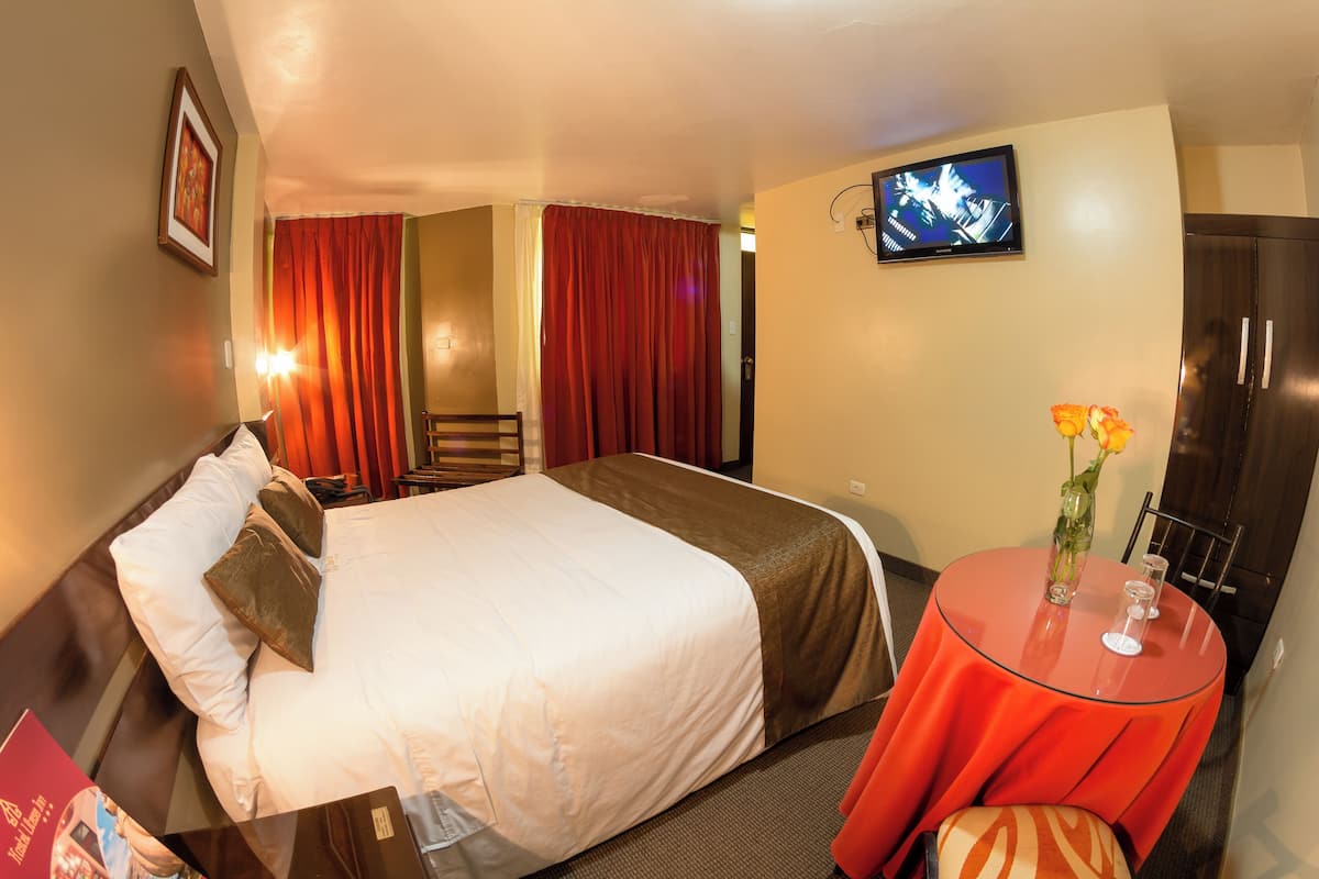 A hotel room with a bed, a TV, a small table with a vase, and a chair.
