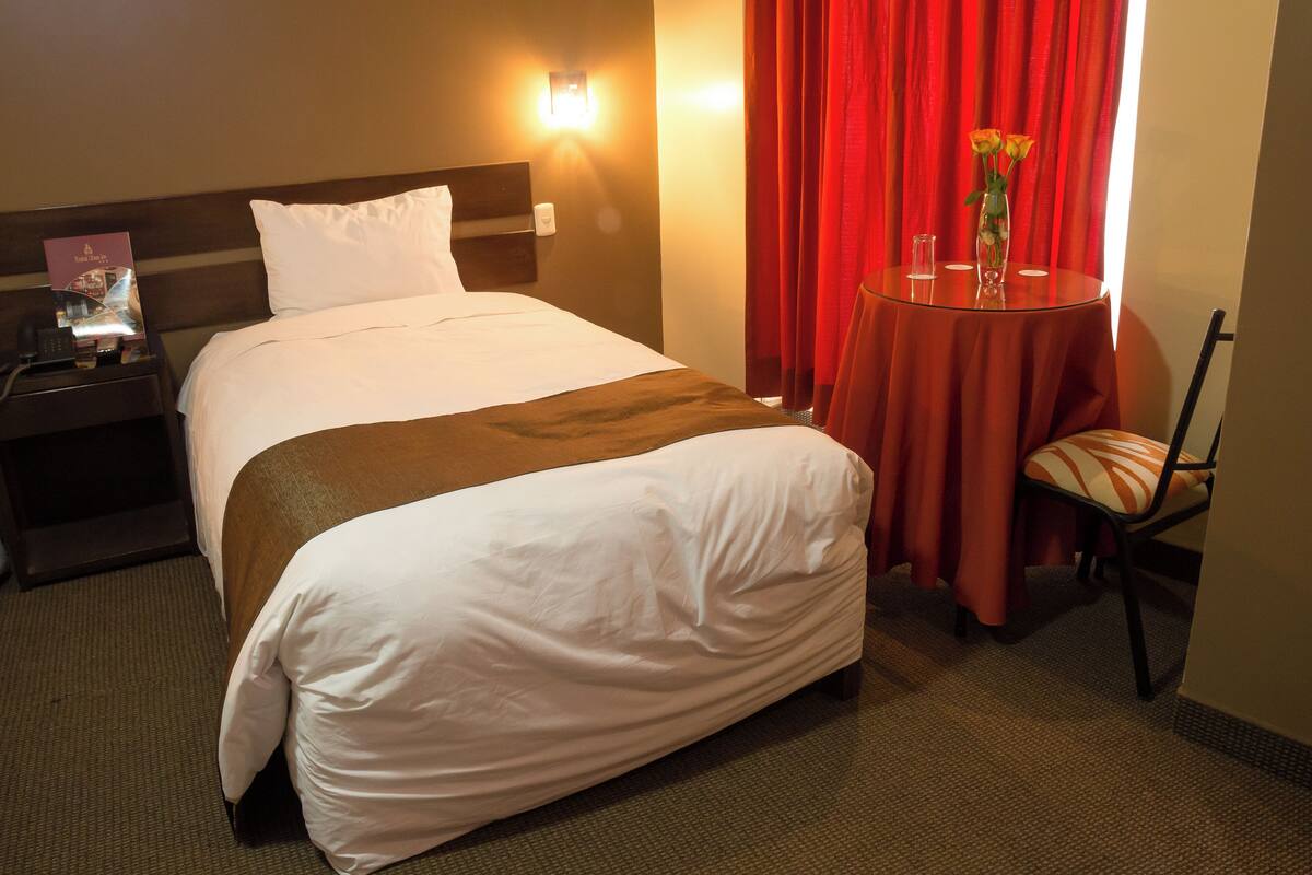 A hotel room with a bed, a nightstand, a table with a vase of flowers, and a chair.