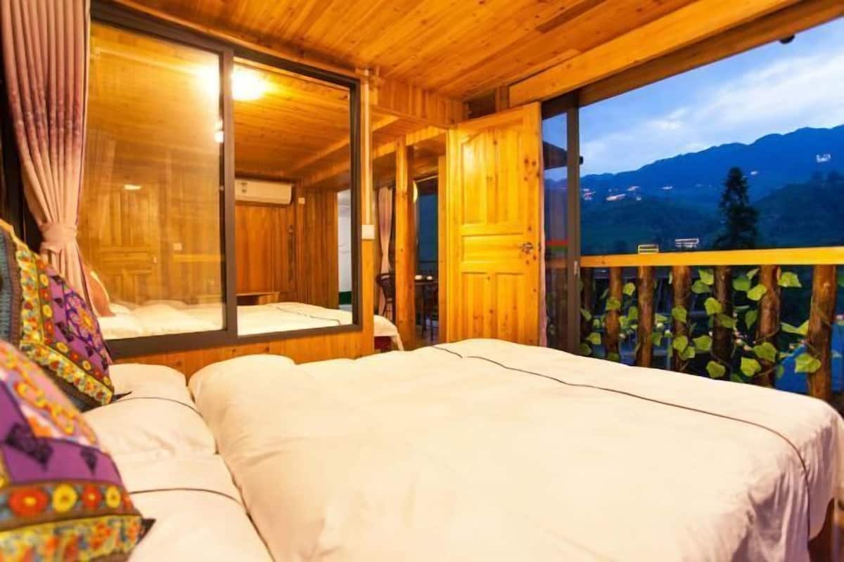 A wooden cabin room with a bed, a window, and a balcony with a view of mountains.