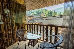 A wooden balcony with two wicker chairs and a glass table, overlooking a village with traditional buildings and mountains. - Xiao Bei Lou (Longsheng)